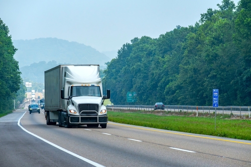 A white semi-truck driving on a highway surrounded by green trees and hills.