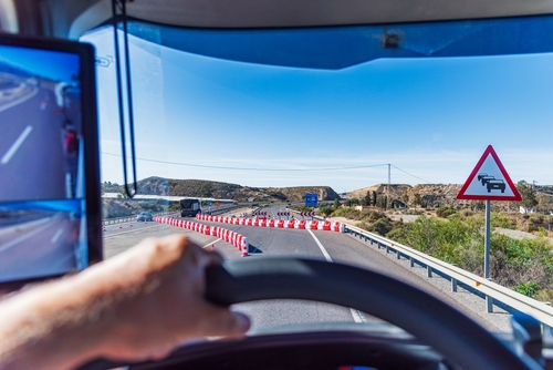 A truck driver's view from inside the cabin, approaching a road construction zone with traffic cones and a warning sign ahead